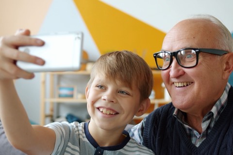 A boy holds a phone at arm's length while his grandfather leans in beside him, both smiling at the screen.