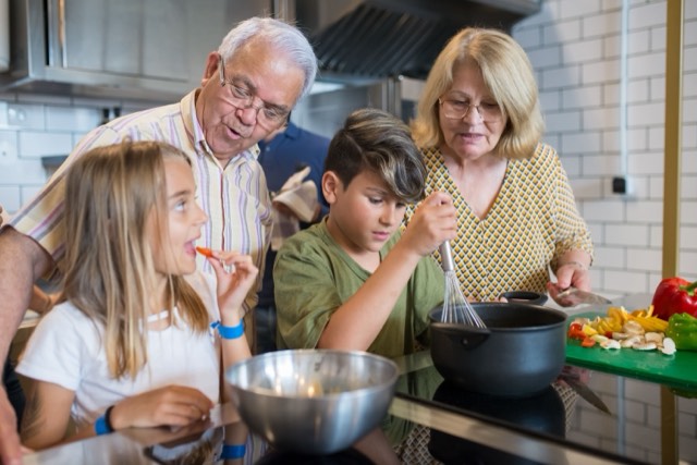 A grandfather and grandmother cook at a kitchen counter with two grandchildren. The boy stirs a pot with a whisk while the girl tastes a slice of tomato.