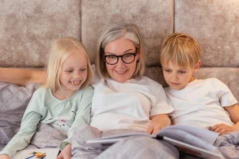 A grandmother in glasses reads a picture book in bed with two grandchildren tucked in on either side of her. The girl is laughing; the boy is watching the page.